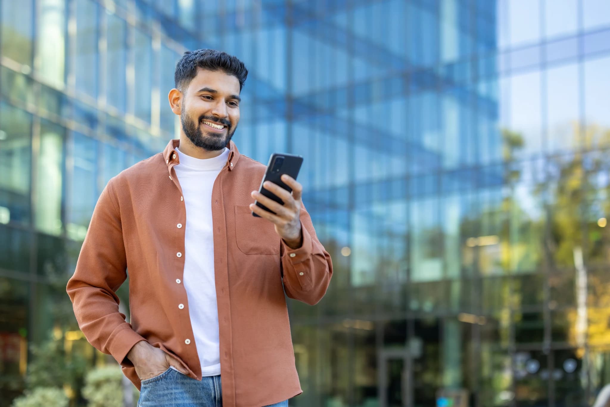 A smiling man looks at his phone while standing outside a modern building during the day.
