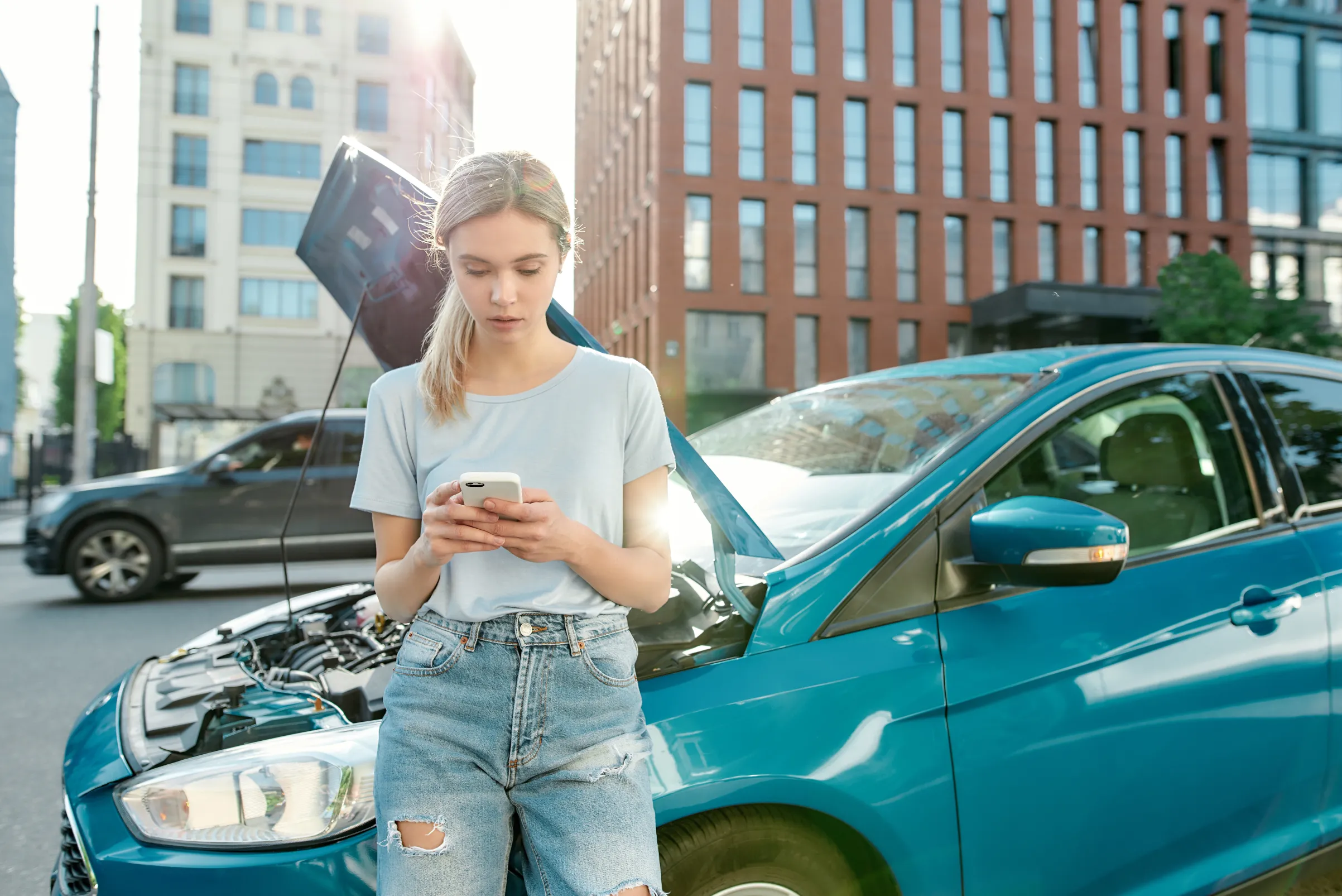 Woman holding phone while standing, leaning on her broken car